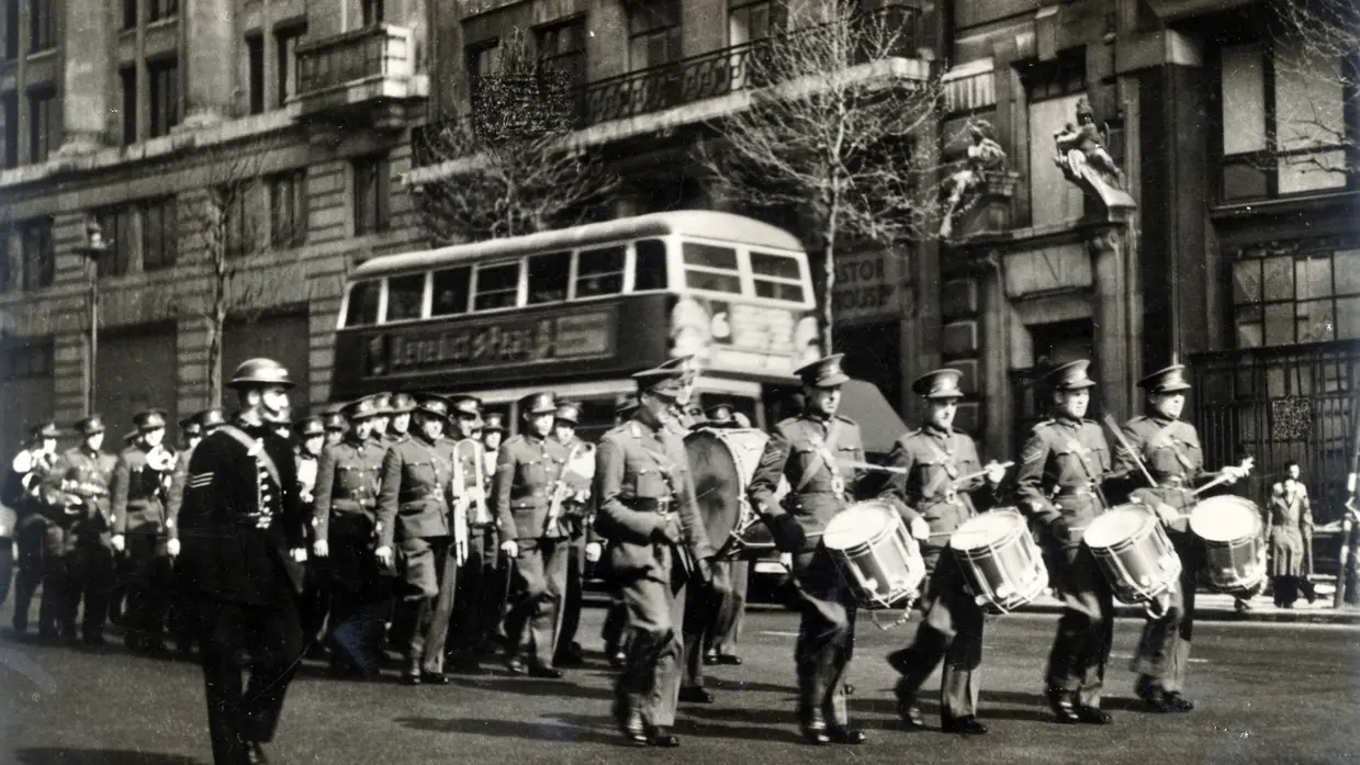 Zwart-witfoto van militair orkest dat speelt op straat met bus op de achtergrond