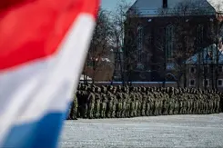 Manschappen in uniform op een rij op de achtergrond en op de voorgrond links een stukje van de Nederlandse vlag.