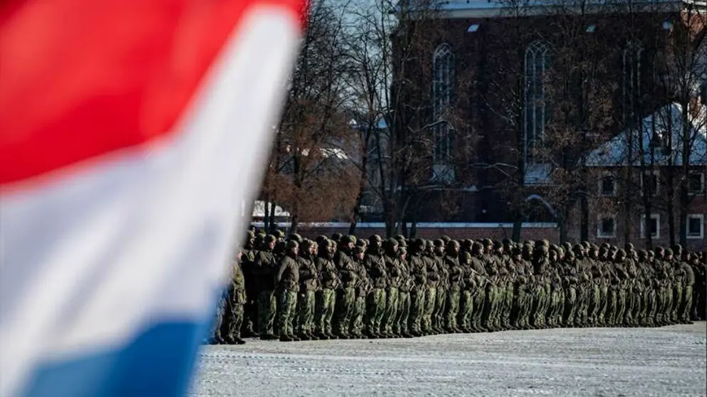 Manschappen in uniform op een rij op de achtergrond en op de voorgrond links een stukje van de Nederlandse vlag.