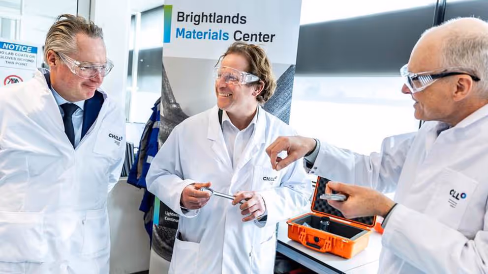 Staatssecretaris Tuinman in witte jas met veiligheidsbril praat bij de Brightlands Chemelot Campus met 2 mannen in witte jassen.