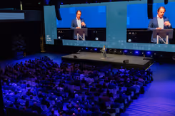 Staatssecretaris Tuinman staat op een podium. Achter hem is hij zichtbaar op 2 schermen. 