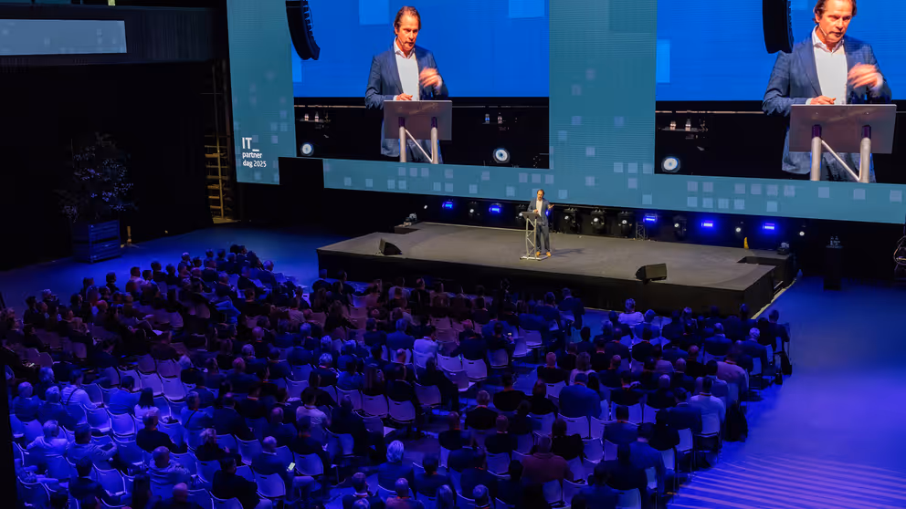 Staatssecretaris Tuinman staat op een podium. Achter hem is hij zichtbaar op 2 schermen. 