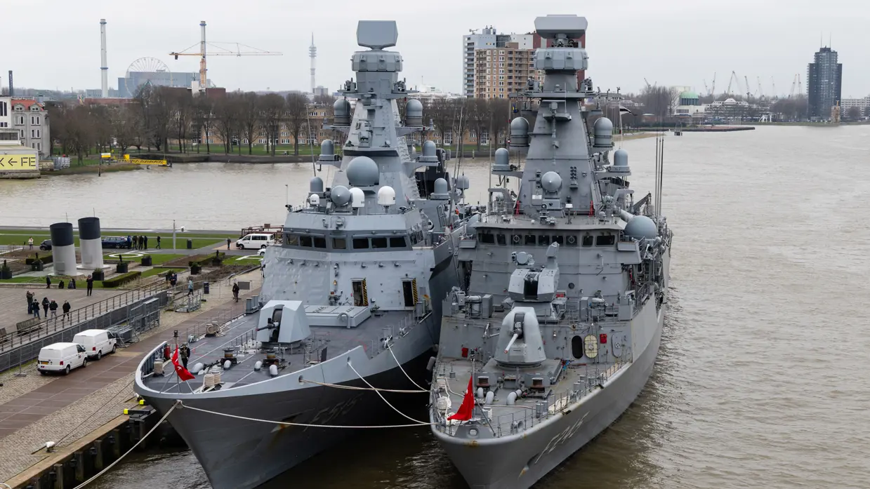 Twee oorlogsschepen liggen in een haven en op een groot schip staan helikopters opgesteld.