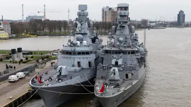 Twee oorlogsschepen liggen in een haven en op een groot schip staan helikopters opgesteld.