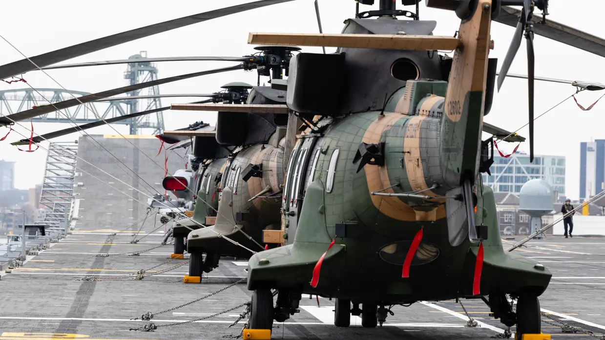 Twee oorlogsschepen liggen in een haven en op een groot schip staan helikopters opgesteld.
