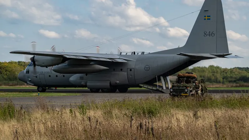 Een transportvliegtuig staat op de baan met de laadklep naar beneden. Dichterbij rijdt een militair voertuig voorbij.