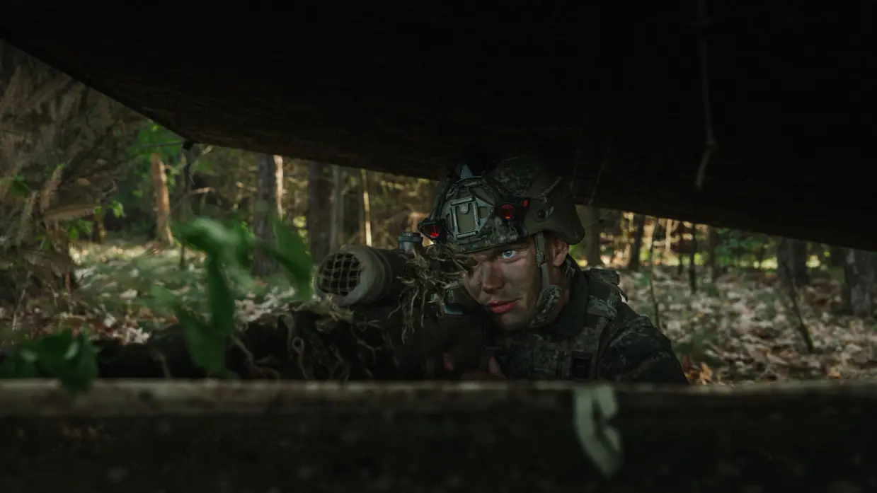 Een gecamoufleerde militair ligt met zijn wapen in stelling onder een schuilnet in een bosgebied tijdens oefening Versatile Guard. 
