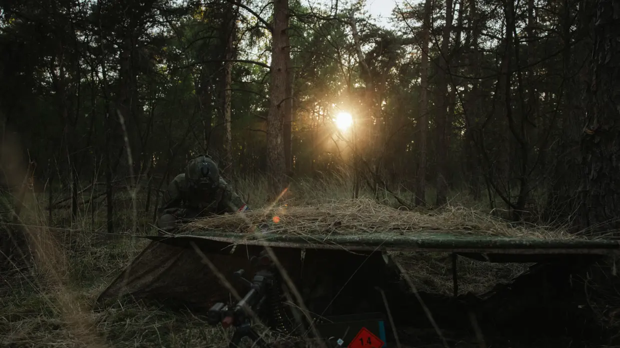 Een militair ligt in stelling naast een gecamoufleerde positie in een bosgebied terwijl de zon opkomt tijdens oefening Versatile Guard.