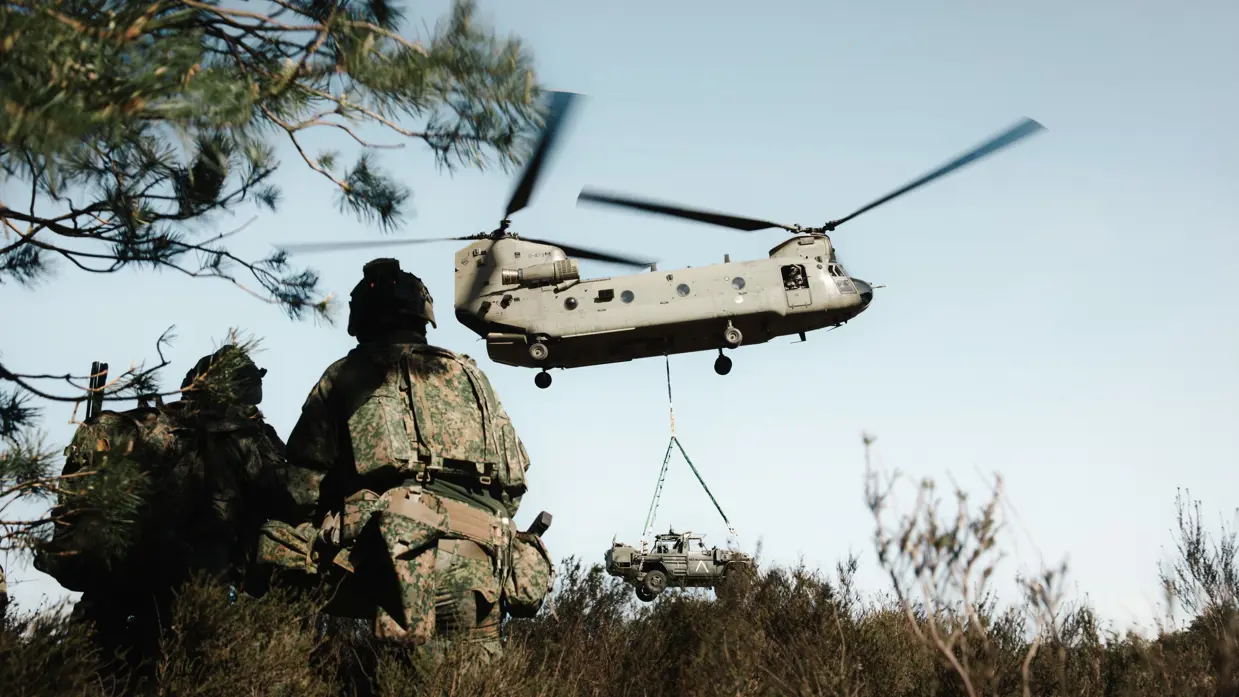 Militairen kijken toe terwijl een Chinook een voertuig onder de romp vervoert tijdens oefening Versatile Guard. 