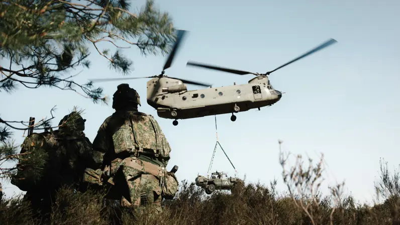 Militairen kijken toe terwijl een Chinook een voertuig onder de romp vervoert tijdens oefening Versatile Guard. 