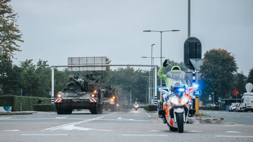 Een militaire colonne rijdt over de weg, daarvoor een motor van de Koninklijke Marechaussee.