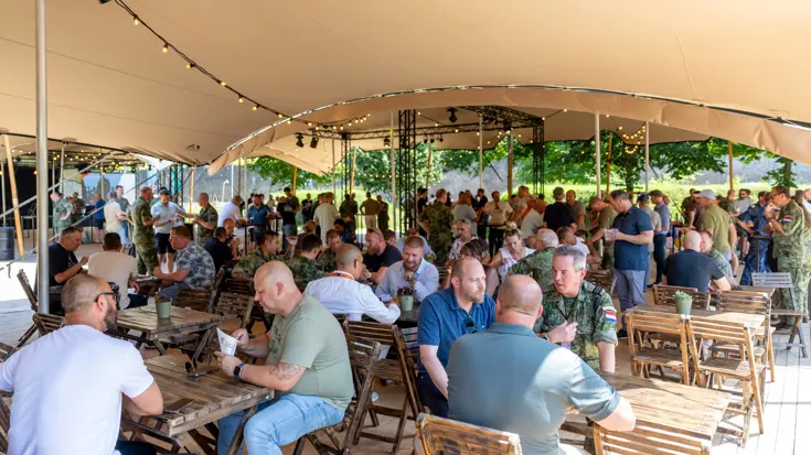 Een overkapping met daaronder kleine houten tafels. Daaraan zitten militairen en burgermedewerkers van COMMIT. Op de achtergrond eenzelfde mix van personen aan statafels. 