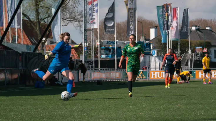 Op een voetbalveld met een tribune aan de zijkant schiet een vrouw in een blauw shirt de bal, terwijl andere speelsters in een groen shirt komen aanrennen. 
