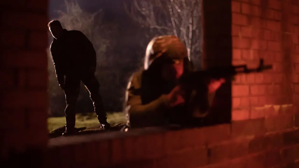 Militair leunt door raam en richt wapen. Een instructeur kijkt op de achtergrond mee.
