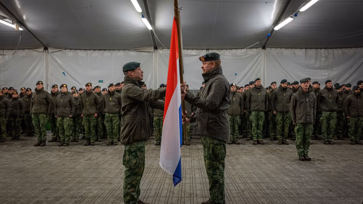 Twee militairen staan tegenover elkaar met een vlag tussen hen in. Op de achtergrond nog meer militairen. 