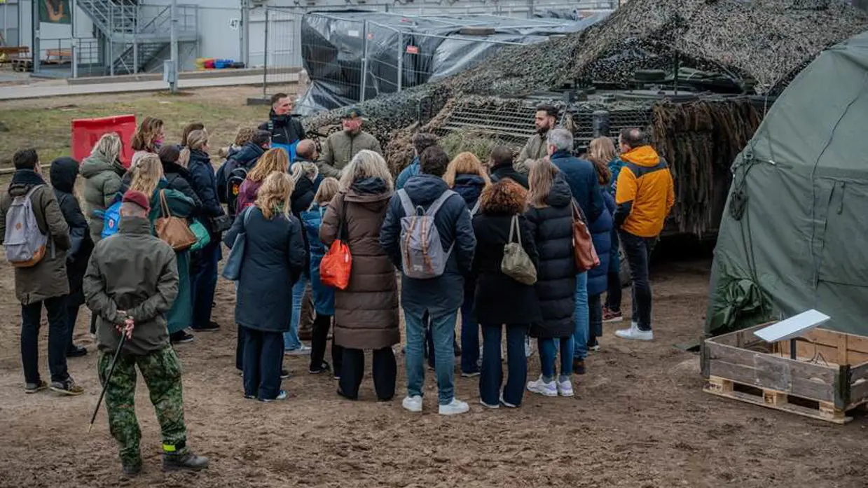 Mensen in burger luisteren naar militair voor de groep. Op de achtergrond militair materieel.