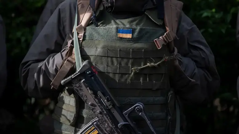 Oekraïense militair in donkere gevechtsuitrusting met een Oekraïense vlag op het kogelwerende vest en een geweer voor de borst.