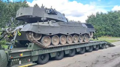 Een tank staat op een groene transporttrailer, omringd door bomen en een heldere lucht met wat wolken.