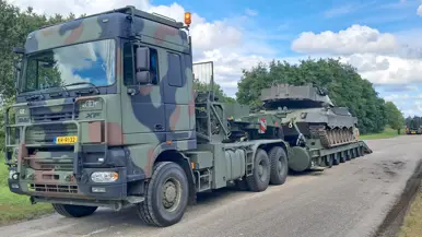 Een camouflage vrachtwagen met Nederlandse kentekenplaat trekt een zware trailer met een tank op een landweg.