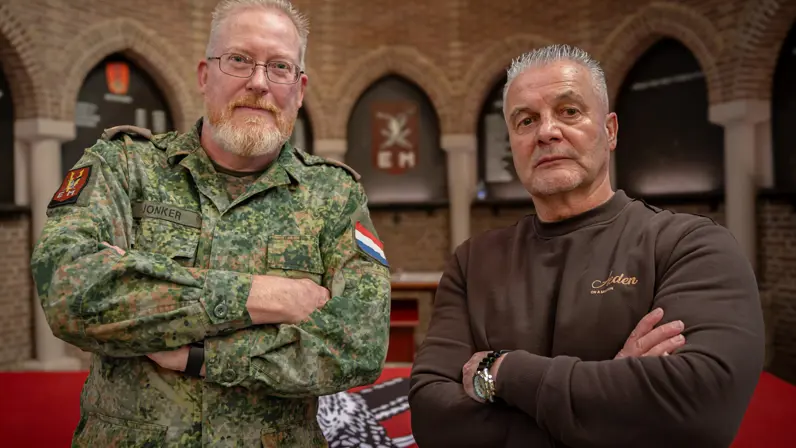 Twee mannen staan met gekruiste armen: links in camouflageuniform, rechts in bruine trui, achter hen een bakstenen muur met bogen.