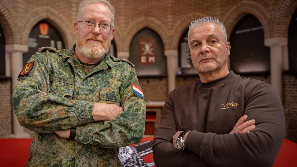 Twee mannen staan met gekruiste armen: links in camouflageuniform, rechts in bruine trui, achter hen een bakstenen muur met bogen.