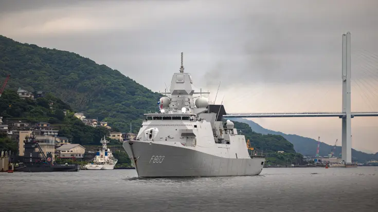 Een varend, grijs marineschip. Op de achtergrond een brug en groene heuvels.